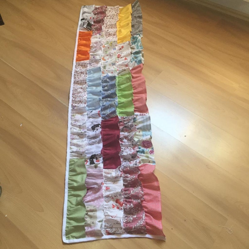 May include: A long, rectangular patchwork quilt runner with a white border. The runner is made of various colorful fabrics, including red, green, blue, yellow, and orange. The fabrics are sewn together in a patchwork pattern, and the runner has a ruffled edge.