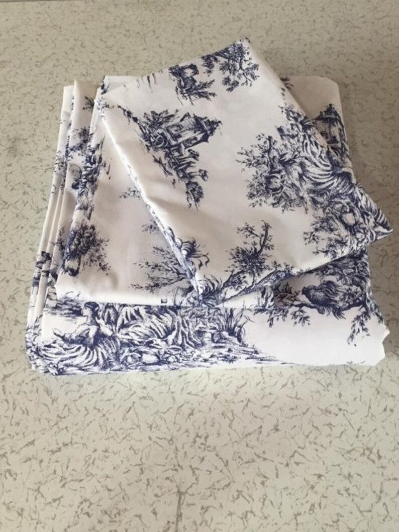 May include: A set of white cotton bedding with a blue toile pattern featuring a repeating design of trees, houses, and birds. The bedding is folded and stacked on a white surface.