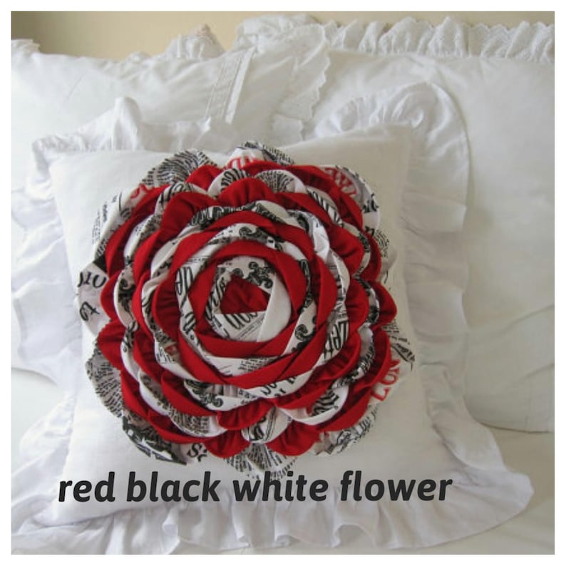 May include: White pillow with a red, black, and white fabric flower in the center. The flower is made of fabric strips sewn together in a circular pattern.