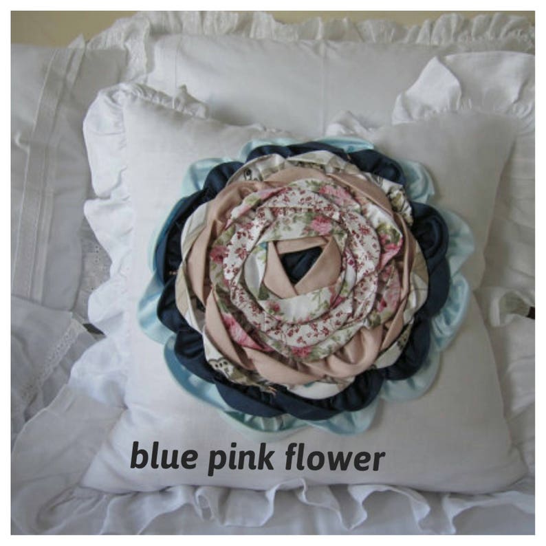 May include: A white pillow with a blue, pink, and white fabric flower in the center. The flower is made of multiple layers of fabric and has a floral pattern.