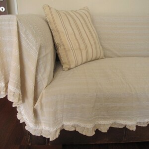 K&ouml;nnte beinhalten: Ein beiges Sofa mit einer wei&szlig;en und beige gestreiften &Uuml;berwurfdecke mit wei&szlig;er Spitzenborte. Das Sofa hat eine braune Lederbasis.