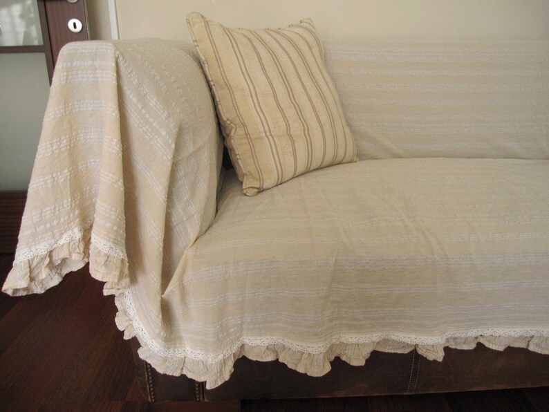 K&ouml;nnte beinhalten: Ein beiges Sofa mit einer wei&szlig;en und beige gestreiften &Uuml;berwurfdecke mit wei&szlig;er Spitzenborte. Das Sofa hat eine braune Lederbasis.