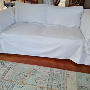 May include: A white sofa with a white slipcover and a white throw pillow. The sofa is sitting on a wooden floor with a blue and gray patterned rug in front of it.