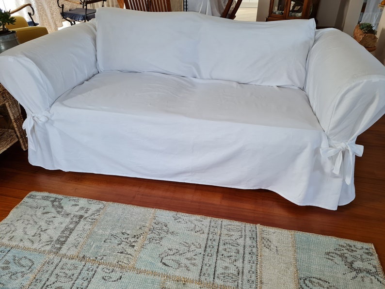 May include: A white sofa with a white slipcover and a white throw pillow. The sofa is sitting on a wooden floor with a blue and gray patterned rug in front of it.