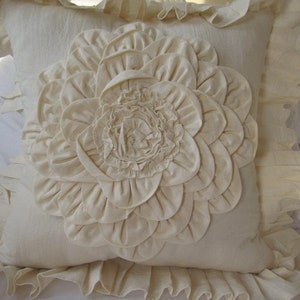 May include: A white pillow with a large, ruffled flower design in the center. The pillow has a ruffled border.