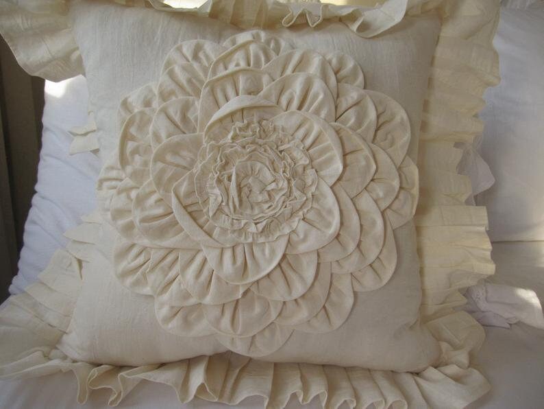 May include: A white pillow with a large, ruffled flower design in the center. The pillow has a ruffled border.