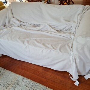 May include: A white sofa cover with ties on the bottom corners. The cover is made of a soft, white fabric and is draped over a sofa.