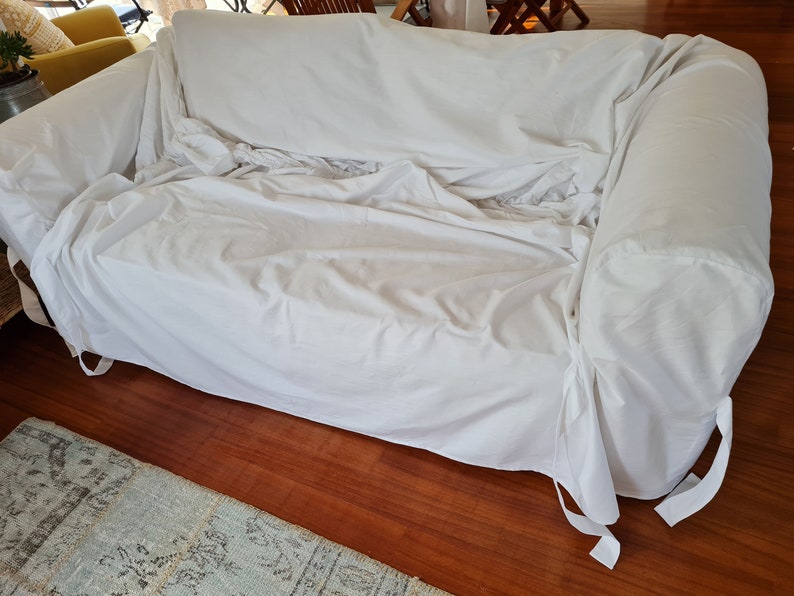 May include: A white sofa cover with ties on the bottom corners. The cover is made of a soft, white fabric and is draped over a sofa.