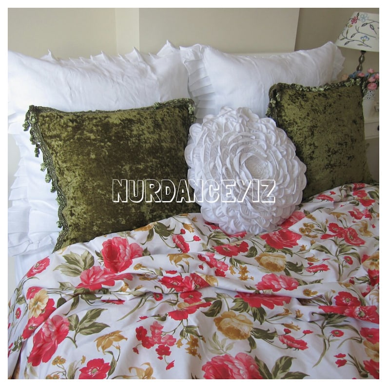 May include: A white bed with a floral patterned duvet cover. The bed is made with white pillows and two green velvet pillows. A large white ruffled pillow is in the center of the bed.