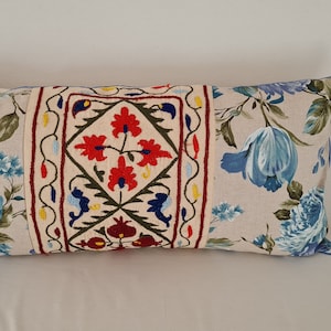 May include: A long rectangular cushion with a beige linen fabric cover. The cushion has a floral pattern in shades of blue and green. The centre of the cushion features a square embroidered panel with a floral design in red, yellow, and green.