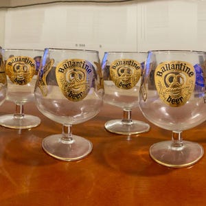 May include: A collection of seven clear glass beer glasses with gold rims. Each glass features a gold and blue label with the words "Ballantine beer" and a pretzel design. The glasses are arranged on a wooden surface.