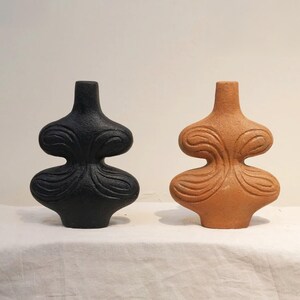 Könnte beinhalten: Zwei dekorative Keramikvasen. Eine ist schwarz, die andere terrakottafarben. Beide haben eine einzigartige, abstrakte Form mit schmalem Hals und breiterem, geschwungenem Körper. Die Vasen stehen auf einer weißen Oberfläche.