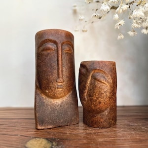 Puede incluir: Dos esculturas de rostros de piedra marrón tallada. La escultura más alta es rectangular con una cara larga y ojos cerrados. La escultura más corta es un cilindro redondeado con un perfil facial. Ambas tienen un aspecto envejecido y texturizado.