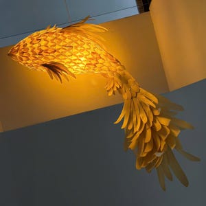 Könnte beinhalten: Eine hängende fischförmige Pendelleuchte, die aus zahlreichen überlappenden gelben schuppenartigen Elementen besteht. Die Struktur strahlt ein warmes, goldenes Licht von innen aus und betont die geschichteten, federartigen Flossen und den Schwanz.