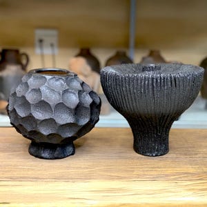 Könnte beinhalten: Zwei schwarze Keramikvasen auf einer Holzoberfläche. Die Vase links hat ein strukturiertes, wabenartiges Muster. Die Vase rechts hat eine ausgestellte Form mit vertikalen Rillen. Beide Vasen haben eine dunkle, matte Oberfläche.