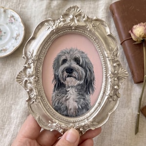 May include: An oval-shaped, ornate silver-toned frame containing a detailed painting of a fluffy, grey and white dog with dark eyes and a black nose, set against a solid light pink background. The frame features intricate floral and shell-like carvings.