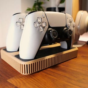 Puede incluir: Un mando de videojuegos blanco y negro descansa sobre una estación de carga beige. El mando tiene un diseño elegante con botones y joysticks visibles. La estación de carga tiene un diseño acanalado.