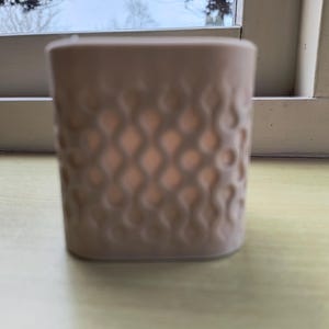 Op de afbeelding: Een beige keramische kandelaar met een gestructureerd, herhalend patroon. De binnenkant gloeit met een warm, zacht licht. De vierkante houder staat op een lichtgele ondergrond.