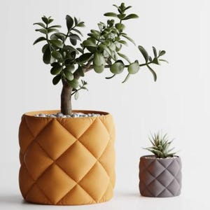 Op de afbeelding: Twee decoratieve plantenbakken met een gewatteerd ruitpatroon. De grotere plantenbak is warm oranje van kleur en bevat een jadeplant. De kleinere plantenbak is grijs en bevat een vetplant. Beide staan op een witte achtergrond.