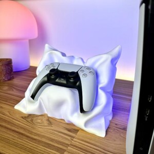 Op de afbeelding: Een witte en zwarte gamecontroller rust op een witte, kussenvormige standaard. De standaard staat op een houten oppervlak, met een paddenstoelvormige lamp op de achtergrond. Een zwart-witte gameconsole staat rechts.