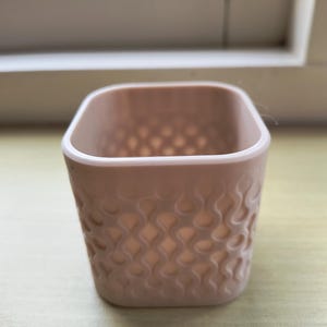 Op de afbeelding: Een kleine, vierkante, lichtroze container met afgeronde hoeken. De buitenkant heeft een herhalend, golvend patroon. De container is leeg en staat op een lichtgekleurd oppervlak, met een raam op de achtergrond.