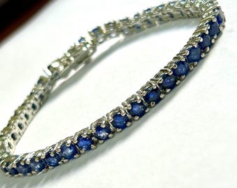 May include: A silver bracelet featuring a series of round, deep blue gemstones. The gemstones are set closely together, creating a continuous line of color. The bracelet has a secure clasp and a delicate, elegant design, suitable for formal or everyday wear.