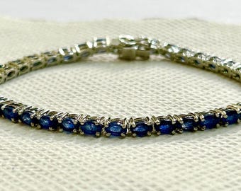 May include: A silver bracelet featuring a line of oval-cut, deep blue gemstones. The gemstones are set closely together, creating a continuous band of color. The bracelet is on a textured, off-white surface.