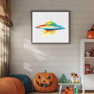 Könnte beinhalten: Ein gerahmtes Aquarellgemälde eines bunten UFOs mit Regenbogen-Design auf weißem Hintergrund. Das Kunstwerk wird an einer weißen Wand ausgestellt, mit einem Kürbis und anderen Dekorationen im Vordergrund.