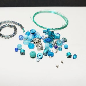May include: A collection of blue and silver beads, including glass, metal, and plastic beads, with a length of teal wire. The beads are perfect for jewelry making.