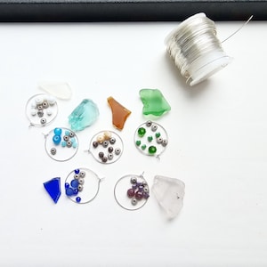 May include: A collection of crafting supplies including a spool of silver wire, five circular wire hoops, and various loose beads in white, blue, green, and purple. Several pieces of smooth, colorful sea glass in white, teal, amber, and green.