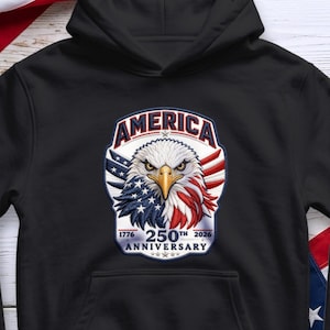 Może przedstawiać: Czarna bluza z kapturem z patriotycznym wzorem. Wzór zawiera orła w czerwono-biało-niebieskiej kolorystyce, napis "AMERICA" oraz tekst "1776 250th 2026 ANNIVERSARY".
