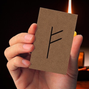 Könnte beinhalten: Eine braune Karte mit einem schwarzen Runensymbol wird in einer Hand gehalten. Die Karte ist rechteckig und die Rune ist ein einfaches, eckiges Design. Der Hintergrund ist dunkel, mit einer Kerzenflamme sichtbar.