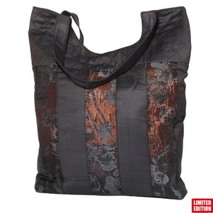 Könnte beinhalten: Eine schwarze Tragetasche mit einem gesteppten Oberteil und einem langen Schulterriemen. Der Hauptteil hat vertikale Bahnen aus dunkelgrauem Stoff mit floralen und strukturierten kupferfarbenen Akzenten. Die Tasche ist eine limitierte Auflage.