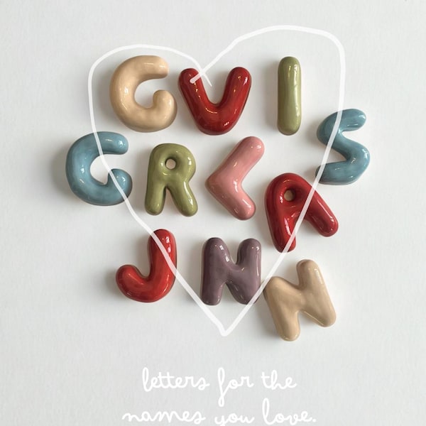 Può includere: Una collezione di undici lettere in ceramica lucida e arrotondata in vari colori, tra cui rosso, blu, verde, beige e viola, disposte a forma di cuore con un contorno bianco disegnato a mano e il testo letters for the names you love.