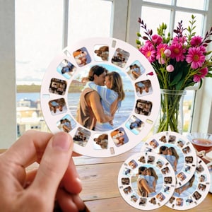 Op de afbeelding: Een witte, op maat gemaakte foto-reelviewer met een centraal beeld van een knuffelend stel, omringd door kleinere afbeeldingen. De viewer wordt voor een raam gehouden met een boeket roze bloemen op de achtergrond. Extra foto-reels liggen op een houten oppervlak.