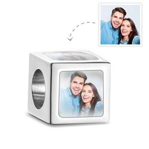 May include: A white, cube-shaped charm with a silver-colored hole. The charm features a photograph of a smiling couple on each side. A smaller image of the couple is shown above the charm.