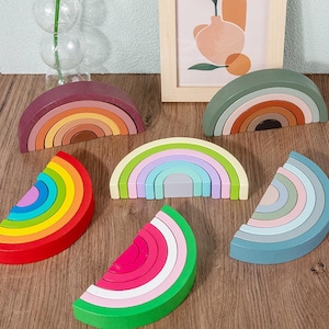 May include: Wooden rainbow stacking toys in various colors, including red, green, and blue. The toys are arranged on a wooden surface. Some are complete rainbows, while others are half-circles or watermelon slices.