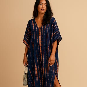 May include: A navy blue caftan dress with vertical orange stripes. The dress has a V-neck and short sleeves. A small, beaded bag is held in the hand. The dress has a slit on the side.