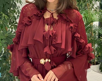 May include: A burgundy dress with a flowing design. The dress has a ruffled bodice, long sleeves, and a matching belt with a gold buckle. The model is wearing a delicate necklace. Green plants are in the background.