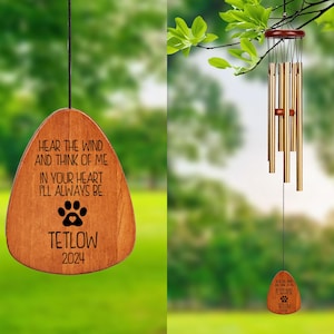 May include: A wooden wind chime with a teardrop-shaped wooden pendant. The pendant has the text "HEAR THE WIND AND THINK OF ME IN YOUR HEART I'LL ALWAYS BE" and the name "TETLOW 2024" with a paw print. The chime has gold-colored tubes.