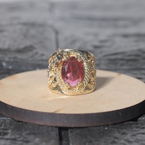 Puede incluir: Un anillo dorado con una gema roja ovalada. El anillo tiene un diseño ornamentado y texturizado alrededor de la gema. El anillo se muestra sobre una pequeña superficie redonda de madera.