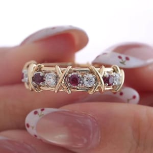 Könnte beinhalten: Ein goldener Ring mit einem abwechselnden Muster aus runden roten Rubinen und klaren weißen Diamanten. Das Metallband ist mit einer Reihe von sich wiederholenden goldenen X-förmigen Kreuzmotiven gestaltet, die jeden der runden Edelsteine trennen.