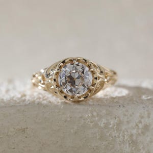 Puede incluir: Primer plano de un anillo de oro con una gema grande, clara y de talla redonda. El anillo tiene un diseño de filigrana ornamentado alrededor de la gema y a lo largo de la banda. El anillo está sobre una superficie texturizada blanquecina.
