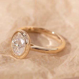 May include: A gold ring with an oval-shaped, clear gemstone. The gemstone is set in a bezel setting with a beaded border. The band is smooth and polished. The ring is displayed on a light brown surface.
