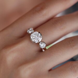 May include: A three-stone ring with a large, round-cut diamond centre stone, with two smaller diamonds either side. The diamonds are set in a white metal band, creating a classic and elegant design. The ring is shown on a person's finger.