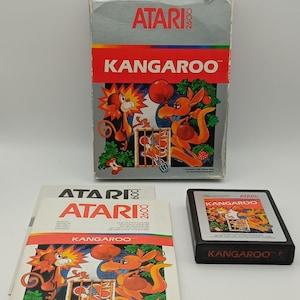 Może przedstawiać: Zestaw gier wideo Atari 2600 dla "Kangaroo". Wyświetlane są pudełko, instrukcja i kartridż. Pudełko zawiera grafikę czerwonego i pomarańczowego kangura. Kartridż jest czarny z kolorową etykietą. Instrukcja jest biała z czerwonym i pomarańczowym tekstem.