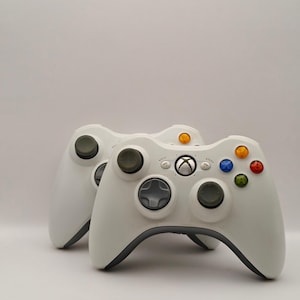 Könnte beinhalten: Zwei weiße Xbox 360-Controller mit grauen Akzenten. Jeder Controller hat ein Steuerkreuz, zwei Analog-Sticks und farbige Aktionstasten. Das Xbox-Logo ist mittig auf dem Controller. Die Controller sind vor einem weißen Hintergrund platziert.