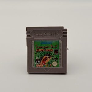 May include: A brown Nintendo Game Boy cartridge featuring the game "Tasmanian Devil: Island Chase." The label is green and red, displaying the game's title and character artwork. The text "Nintendo GAME BOY" is printed on the top of the cartridge.