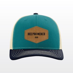 May include: A teal and navy blue baseball cap with a beige mesh back. The front features a tan hexagonal patch with the black text BEE PRO MERCH and the number 2026. The navy blue brim has white stitching details along the curved edge.