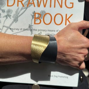 Pode incluir: Uma pulseira de punho bicolor em ouro e preto. A pulseira é usada em um pulso, com um livro intitulado "DRAWING BOOK" ao fundo.